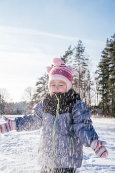 Little girl 3-4 years old in winter overalls, hat and mittens, stands outdoors against against blue sky and fir trees and throws snow over her laughing. Winter holidays, children outdoors. Vertical
