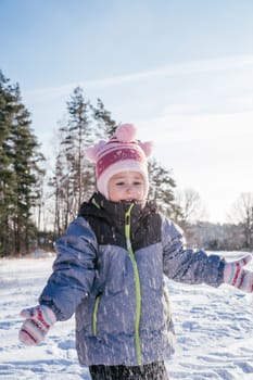Little girl 3-4 years old in winter overalls, hat and mittens, stands outdoors against against blue sky and fir trees and throws snow over her laughing. Winter holidays, children outdoors. Vertical
