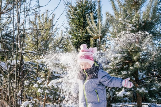 Little girl 3-4 years old in winter clothes overalls, hat and mittens, stands outdoors against snow-covered pines and fir trees and throws snow over her, laughing. Winter holidays, children outdoors
