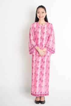 Full body portrait of happy Southeast Asian girl in pink batik dress standing on plain background.