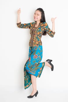 Full length portrait of Southeast Asian woman in batik dress thumbs up, cheering and jumping around on plain background.