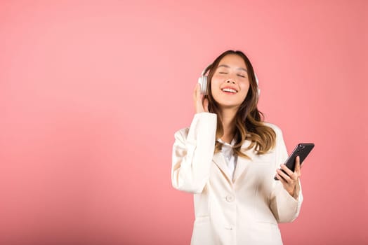 Happy female using mobile phone listens loud song, Portrait of Asian beautiful young woman has fun close eyes listening music with headphones in smartphone app, studio shot isolated on pink background