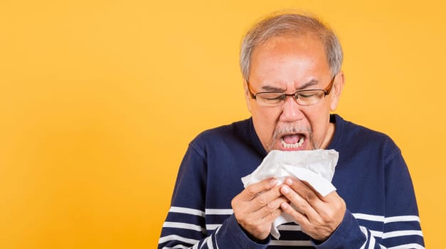 Asian elder man cold having flu and sneezing from sickness virus problem use tissue studio shot isolated on yellow background, Portrait senior old man blowing nose with tissue, pensioner unwell