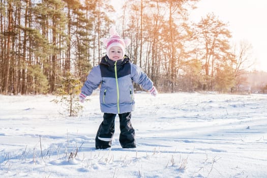 Little girl 3-4 years old in winter clothes overalls, hat and mittens jumps on snow and laughs against snow-covered pines and fir trees on sunny day. Winter holidays, children playing outdoors