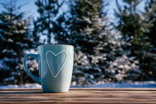 Turquoise blue cup with heart rising steam on wooden table against backdrop of pines and firs with snow on cold winter day. Hot warm drink in frosty weather