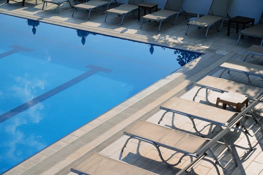 top view closeup detail of sunlounger chairs near an outdoor pool and cloudy sky reflecting in the blue water
