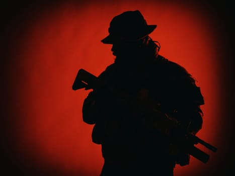 Commando shooter, army special forces rifleman in battle uniform, boonie hat, ammunition in load carrier, camouflaged with paint face, armed assault rifle, half length solhouette portrait on red background