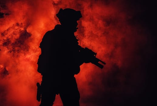 Silhouette of modern infantry soldier, elite army fighter in tactical ammunition and helmet, standing with assault service rifle in hands in the fire and smoke