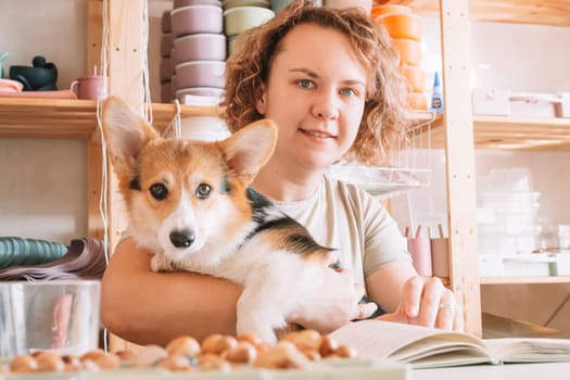 Curly haired glad, cheerful businesswoman, owner with dog corgi sitting, relaxing and reading book at workplace indoors. Small business, silicone dishes for babies. Home comfortable office, store
