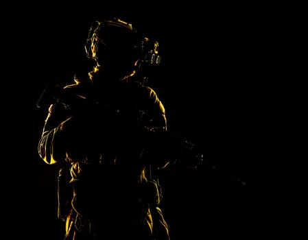 Army special forces elite soldier with hidden behind mask and glasses face, battle helmet, tactical radio headset, standing with assault rifle equipped silencer in darkness, contour shot