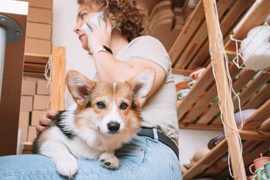 Smiling concentrated businesswoman talking smartphone, holding cute calm dog corgi in workshop. Set of silicone baby dishes near wall on shelves. Home office, taking orders in comfortable atmosphere