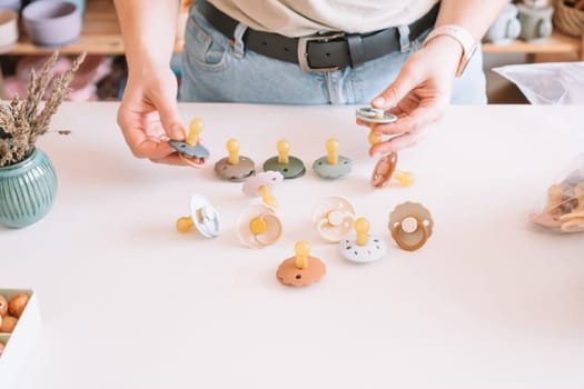 Cropped business woman hands, making and design craft silicone pacifiers for newborn babies on white desk. Small business, store. Choosing soft material for kids. Natural ecological accessories.
