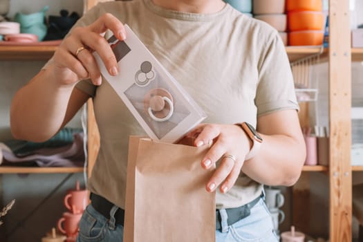 Cropped photo of businesswoman hands packing, preparing parcel for delivery package to buyer. Woman puts pacifier in kraft paper bag. Small business owner, workshop, startup. Close up