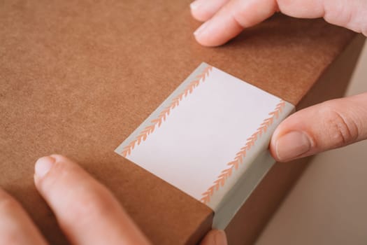 Cropped photo of female hands packing, preparing with scotch tape, parcel for delivery package to buyer. Woman sticks sticker to kraft cardboard box. Small business owner, workshop, startup. Close up