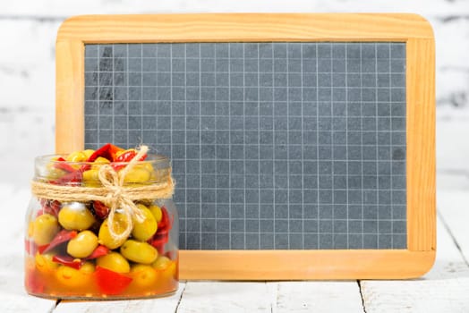 glass jar with olives and peppers with blackboard on white table