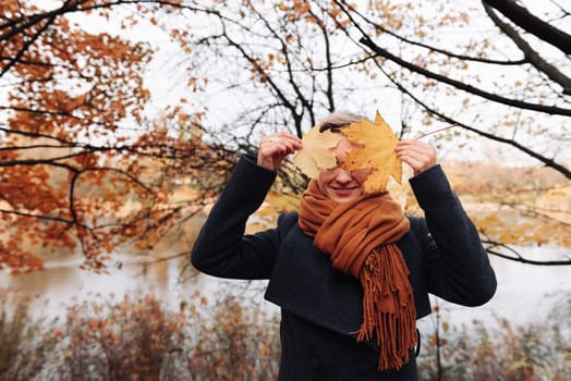 A woman in a coat and scarf in an autumn park covers her face with maple leaves.
