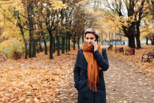Short-haired blonde communicates on the phone walking in the autumn park in a coat and scarf.