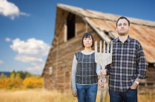 Biracial Chinese and Caucasian Young Couple Holding Wooden Pitch Fork In Front of Rustic Barn In The Country.