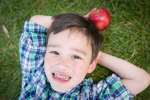 Mixed Race Chinese and Caucasian Young Boy With Apple Relaxing On His Back Outside On The Grass.