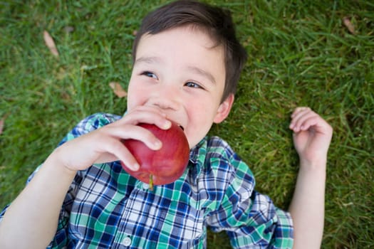 Mixed Race Chinese and Caucasian Young Boy With Apple Relaxing On His Back Outside On The Grass.