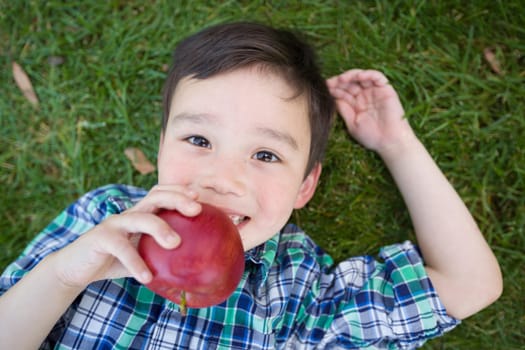 Mixed Race Chinese and Caucasian Young Boy With Apple Relaxing On His Back Outside On The Grass.