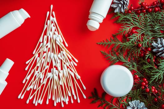 Wooden cotton swabs laid out in shape of Christmas tree with cosmetics jars, roll-on deodorant and spruce branches, cones, red berries on red background. Flat lay