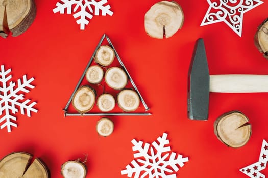 Long nails and wood cuts or branch slabs, laid out in shape of Christmas tree with hammer and wooden snowflakes on red background. Flat lay