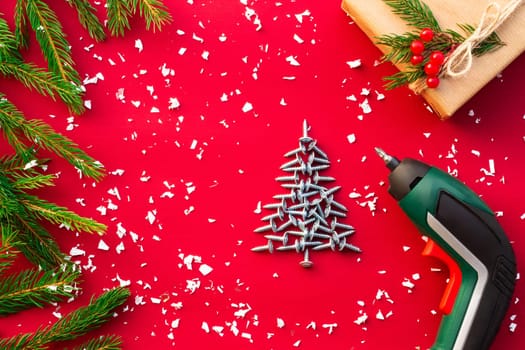Christmas tree made of self-tapping screws, screwdriver, gift in craft paper with branches of a Christmas tree and red berries on a red background. Flat lay. Copy space.