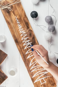 Womans hand draws Christmas tree on decorative board made of burnt wood with brush of white paint. Christmas DIY. Vertical