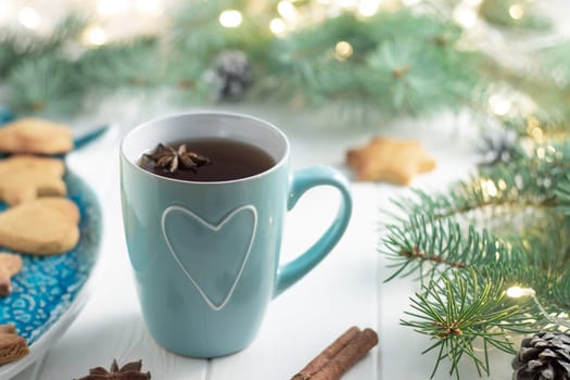 Mint cup of tea with heart, gingerbread, anise, cinnamon next to green branches of Christmas tree with LED light bulbs garland on a white wooden background. Merry Christmas and Happy New Year concept