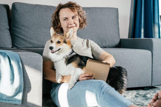 Curly haired glad, cheerful barefoot woman with dog corgi sitting, relaxing and reading book on carpet in comfortable living room near sofa. Calm and cozy atmosphere. Weekend vacation