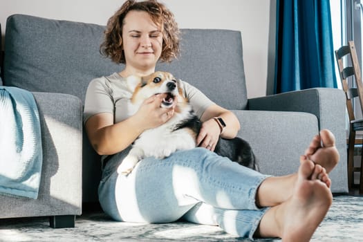 Curly haired glad, cheerful barefoot woman with dog corgi. Obedience training, feeding of pet, sitting, relaxing and playing on floor in comfortable living room near sofa. Weekend leisure activity