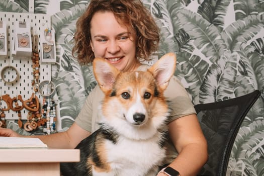 Curly haired cheerful woman, owner with dog corgi sitting, in computer chair near laptop at workplace indoors. Small business, rattles, teethers for babies. Home office, online store, marketplace.