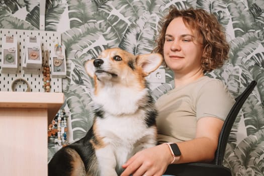 Curly haired cheerful woman, owner with dog corgi sitting, in computer chair near laptop at workplace indoors. Small business, rattles, teethers for babies. Home office, online store, marketplace.