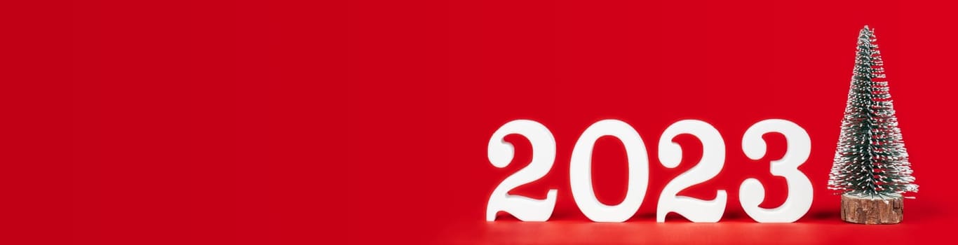 Happy New Year 2023. Bright red long rectangular banner with white wooden numbers 2023 stand with toy Christmas tree. Merry Christmas. Copy space. Place for text