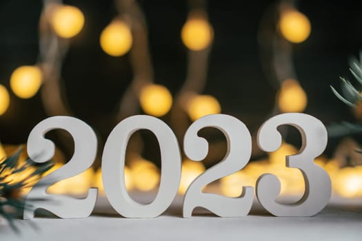 Happy New Year 2023. White wooden numbers 2023 stand on white surface with Christmas tree branches against background of garland light bulbs. Merry Christmas. Copy space