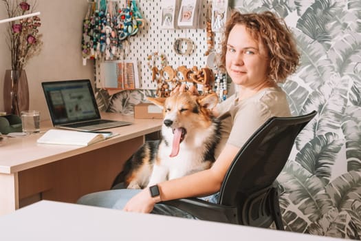 Curly haired cheerful woman, owner with dog corgi sitting, in computer chair near laptop at workplace indoors. Small business, rattles, teethers for babies. Home office, online store, marketplace.