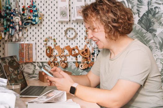 Smiling concentrated businesswoman talking smartphone, working on laptop, taking order. Baby healthcare accessories, silicone eco rattles, pacifiers, teethers hanging on white wall. Work at home