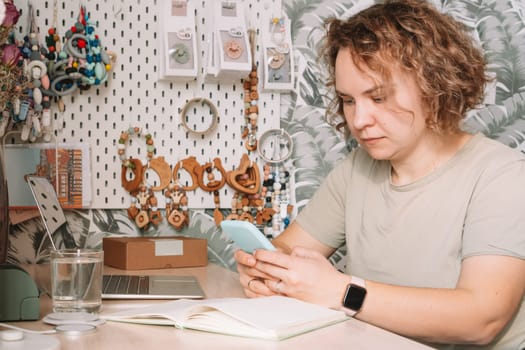 Smiling concentrated business woman talking smartphone, working on laptop in workshop. Baby healthcare accessories, silicone eco rattles, teethers, pacifiers, hanging on white wall. Home office