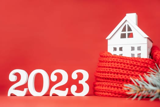 Happy New Year 2023. White wooden numbers 2023 stand with white toy house wrapped in red scarf and Christmas tree branch on red background. Merry Christmas. Copy space