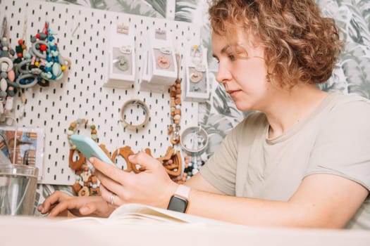 Smiling concentrated businesswoman talking smartphone, working on laptop, taking order. Baby healthcare accessories, silicone eco rattles, pacifiers, teethers hanging on white wall. Work at home