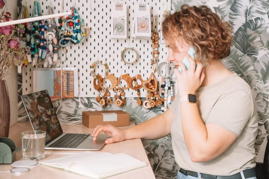 Smiling concentrated business woman talking smartphone, working on laptop in workshop. Baby healthcare accessories, silicone eco rattles, teethers, pacifiers, hanging on white wall. Home office