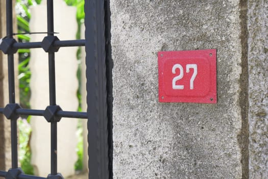Close-up of a red house number 27 mounted on a wall next to an iron gate.