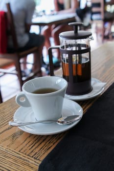 White cup of tea with glass teapot behind on wood table in small cozy cafe