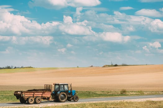 Tractor With Cart And Compost Moving On Country Road In Spring Season. Agricultural Landscape.