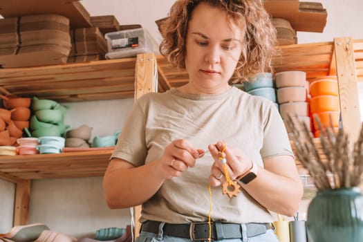 Woman making natural safe accessories of wood on desk, eco style. Stringing beads. Teether toy with trinket rattle decoration. Handcrafting work of toys for baby teeth chewing. Step by step. Kid store