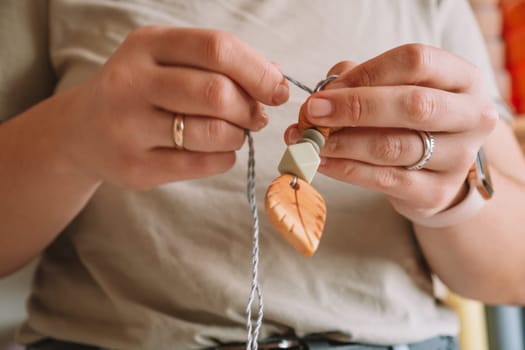 Cropped woman making natural safe accessories of wood on desk, eco style. String beads. Teether toy with trinket rattle decor. Crafting work of toys for baby teeth chewing, hygiene. Home hobby, diy