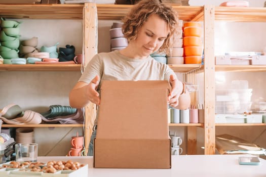 Smiling blond curly woman packing and wrapping shipment, silicone baby dishes in cardboard delivery box. Brown wooden beads in container nearby. Workshop store, small business