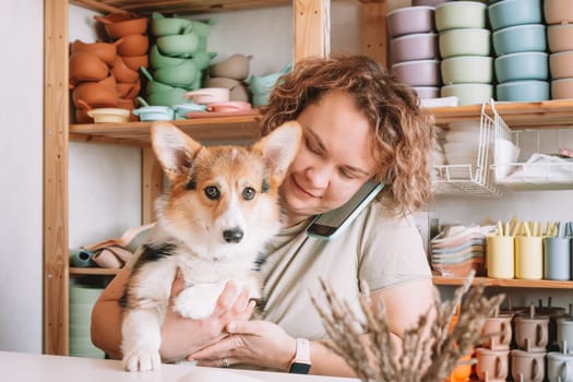 Smiling concentrated businesswoman talking smartphone, holding cute calm dog corgi in workshop. Set of silicone baby dishes near wall on shelves. Home office, taking orders in comfortable atmosphere