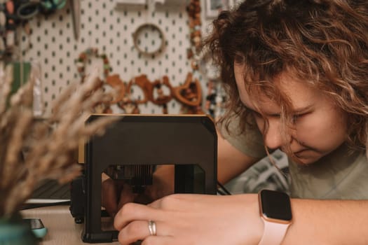Curly haired woman holds item in desktop laser wood burning machine to inscribe name on surface of childrens wooden toy. Home production of teethers and rattles for babies